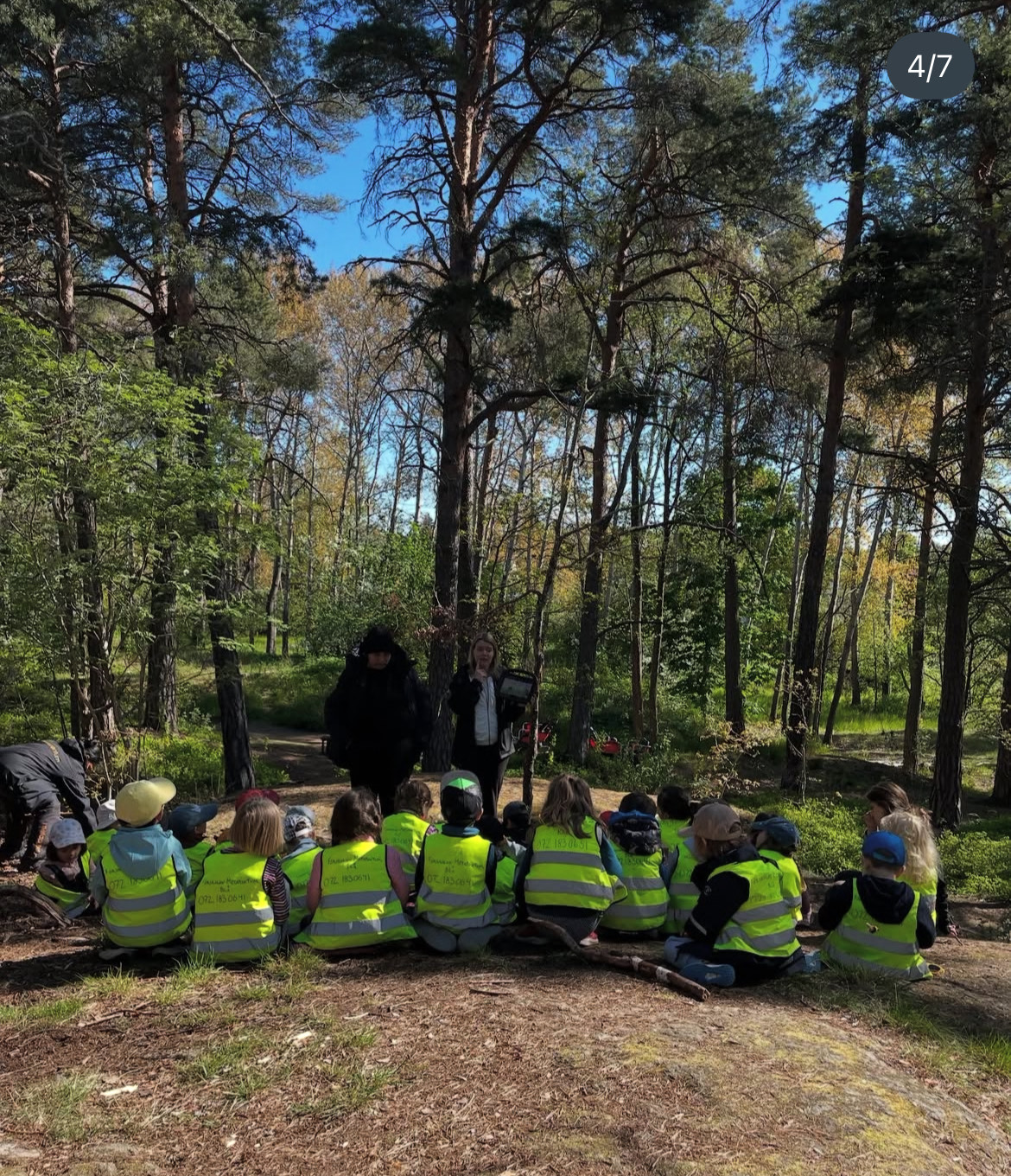 En bild av barn i reflexvästar som sitter samlade på marken framför sina klassföreståndare i skogen